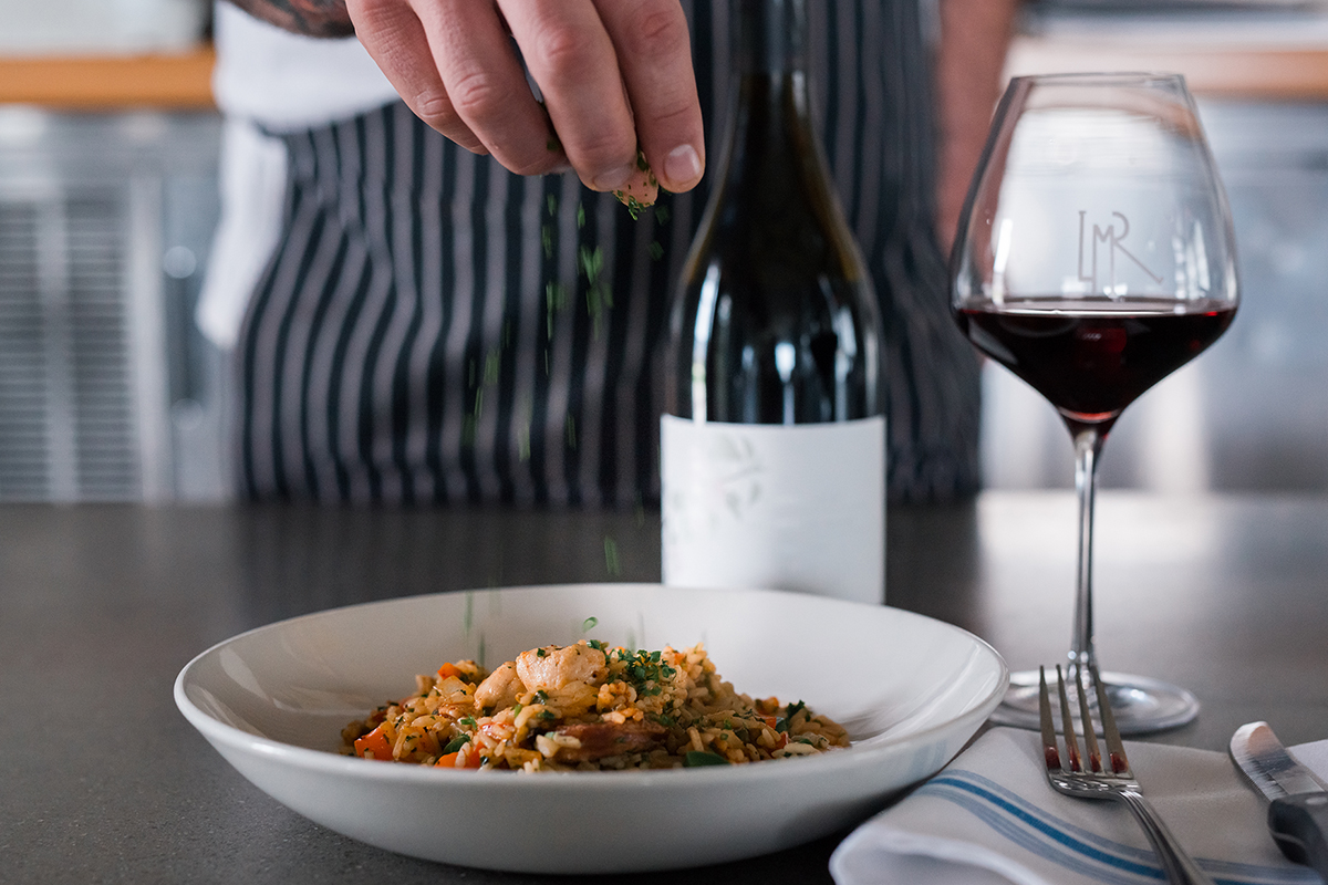 Chef sprinkling herbs on a finished plate of food next to a bottle of Long Meadow Ranch wine