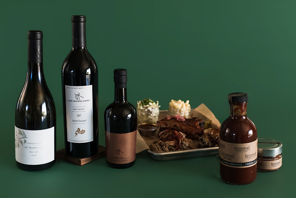 Bottles of Long Meadow Ranch Pinot Gris, Cabernet Sauvignon and BBQ sauce next to a plate of ribs, sides and sauces