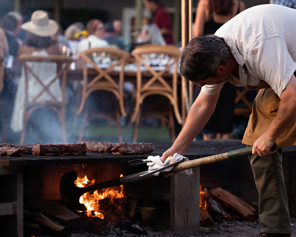 Chef stoking the fire underneath the grill filled with meat